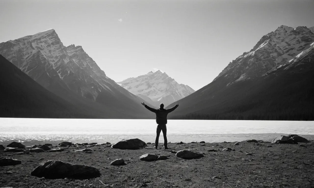 A black and white photograph captures a solitary figure standing at the base of a towering mountain range, their outstretched arms reaching towards the heavens, questioning the height of God.