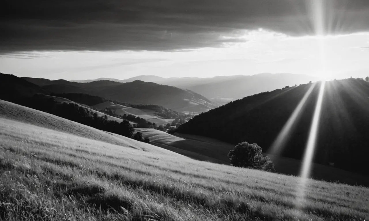 A black and white photo capturing a vibrant sunset over a hilly landscape, where rays of light burst through the clouds, creating an ethereal glow, symbolizing the divine attire of God.