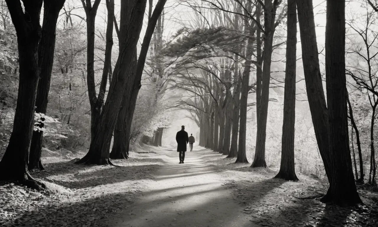 A black and white photo capturing a solitary figure walking along a serene path, surrounded by towering trees, symbolizing the act of humbly journeying with God in peaceful solitude.