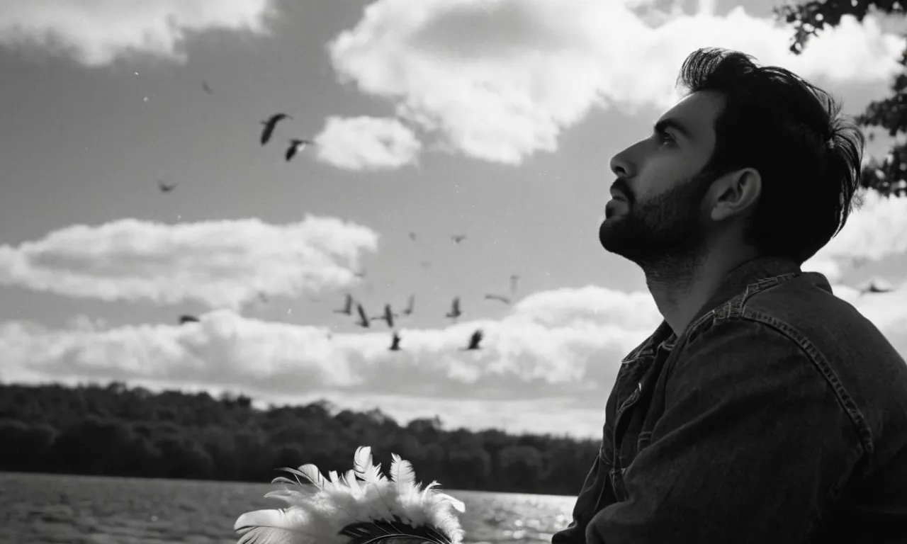 A black and white close-up shot of a solitary figure sitting on a bench, looking up at the sky with a contemplative expression, surrounded by feathers gently falling from above.